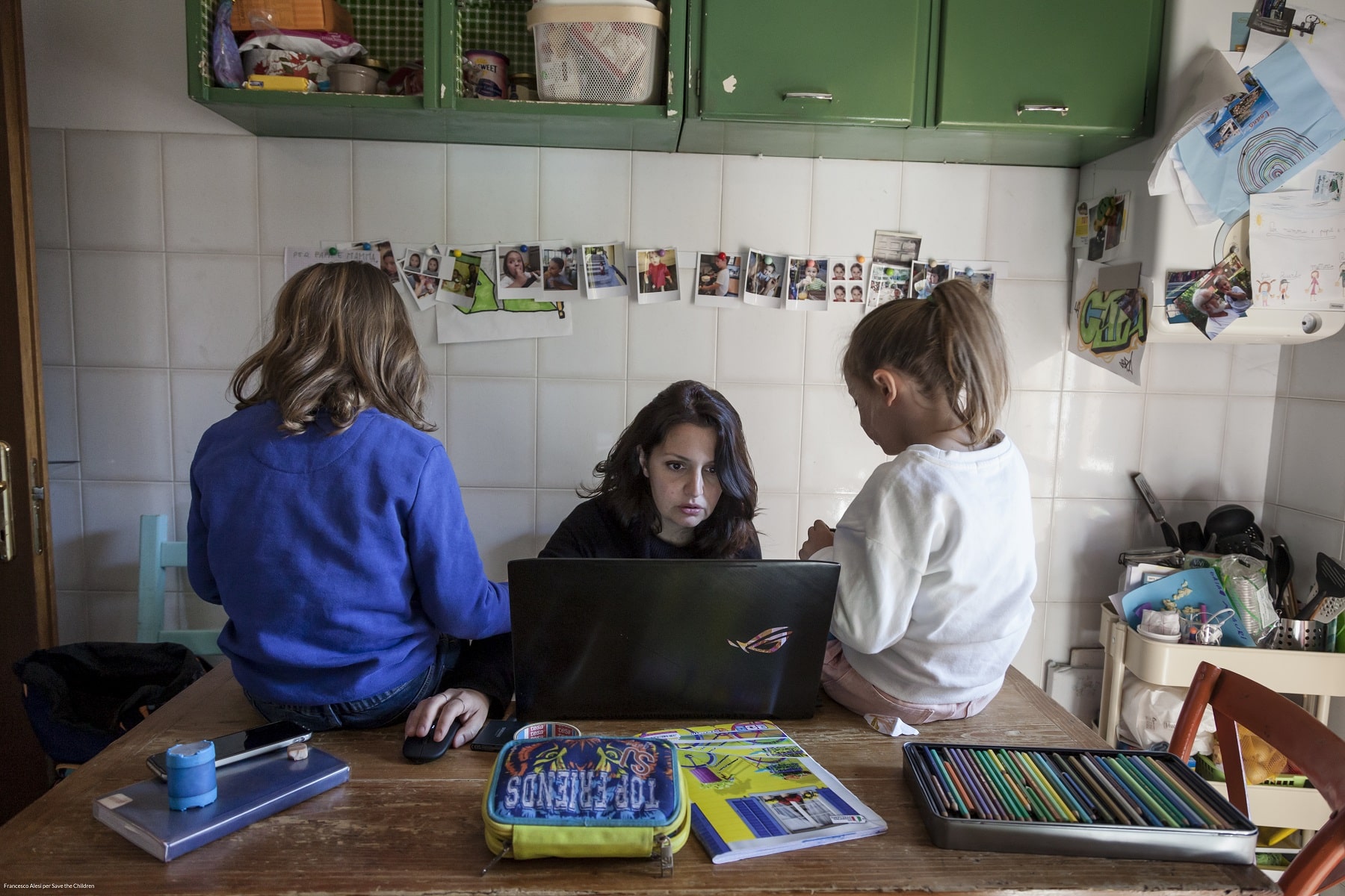 mamma che lavora al pc mentre le due figlie sono sedute sul tavolo accanto a lei 