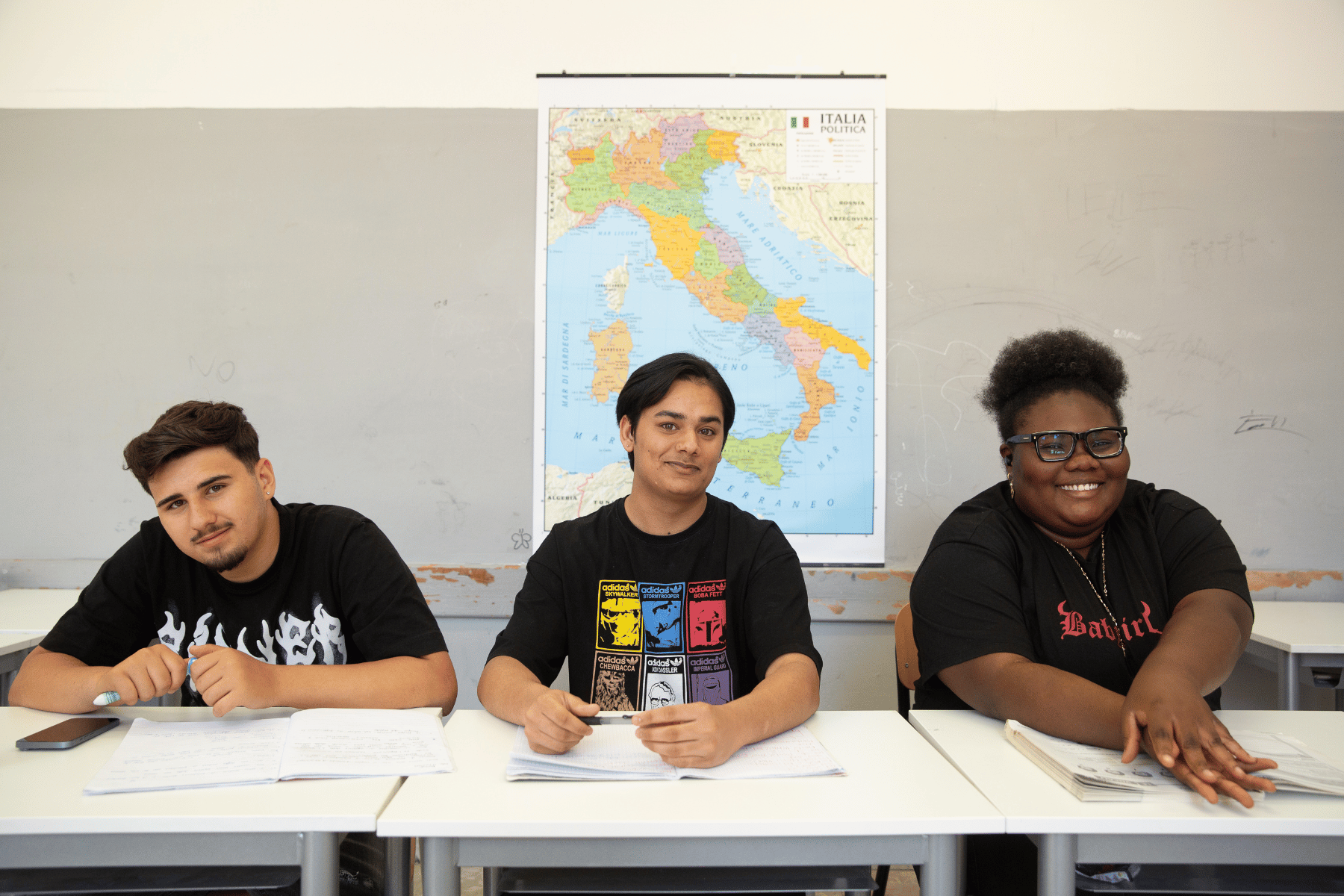 Studentessa e studenti con background migratorio sorridenti su banco di scuola con dietro la cartina d'Italia