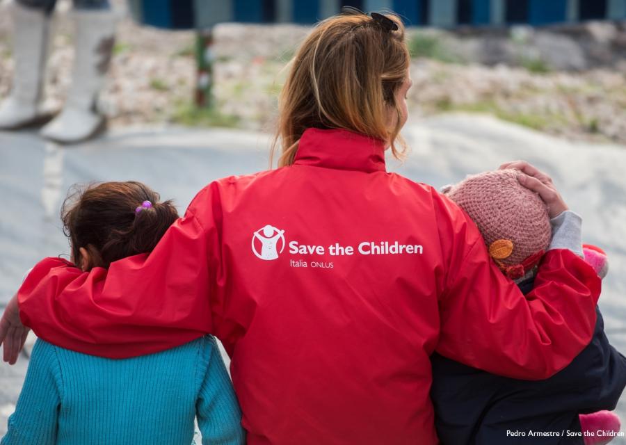 operatrice save the children di spalle con giubbotto rosso tiene sottobraccio due bambini
