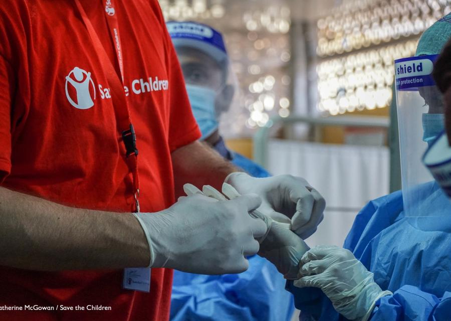 dettaglio su mani con guanti di un operatore save the children con maglietta rossa e di una dottoressa con camice blu e protezioni per coronavirus