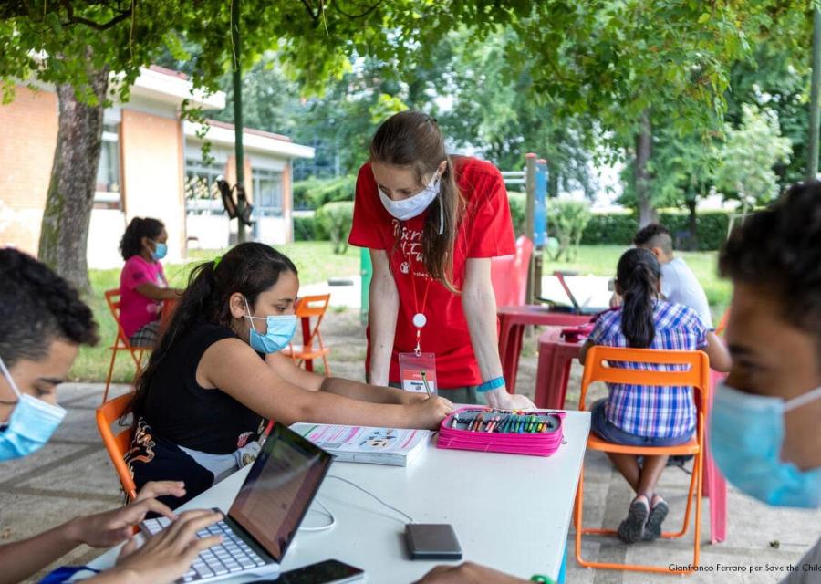operatrice svae the children con dei bambini, seduti a un tavolo esterno mentre fanno attività educative, tutti con mascherina per proteggersi dal covid