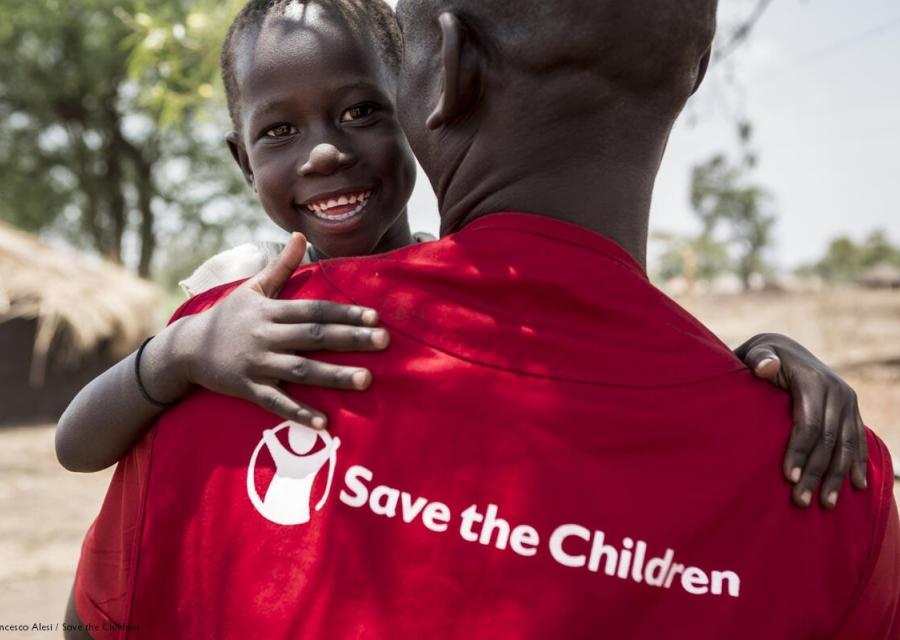 operatore save the children di spalle con maglietta rossa e logo con in braccio un bambino sorridente