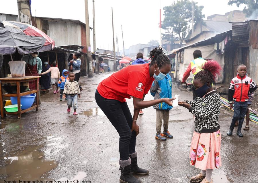 operatrice save the children con maglietta rossa e mascherina mentre assiste una bambina anch'essa con mascherina in una strada