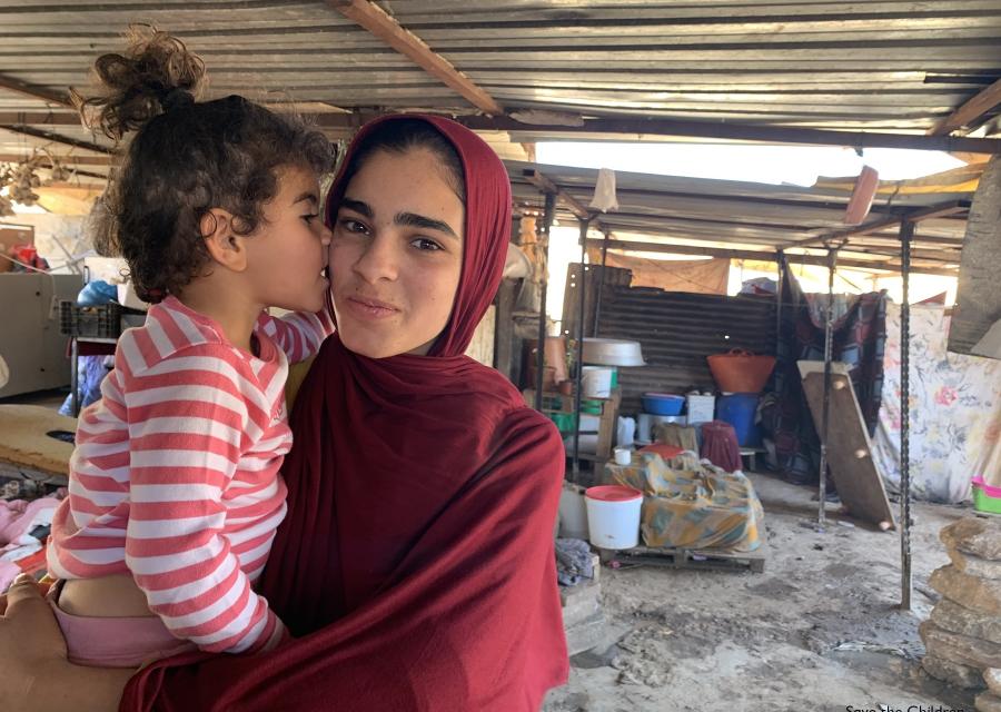 ragazza con velo rosso tiene in braccio una bimba con maglietta a righe bianca e rossa mentre si trovano un un allestimento temporaneo nei territori palestinesi occupati