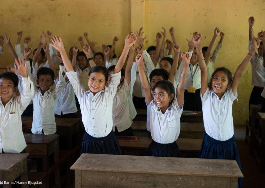 bambine e bambini tra i banchi di scuola con le mani in alto sorridenti 