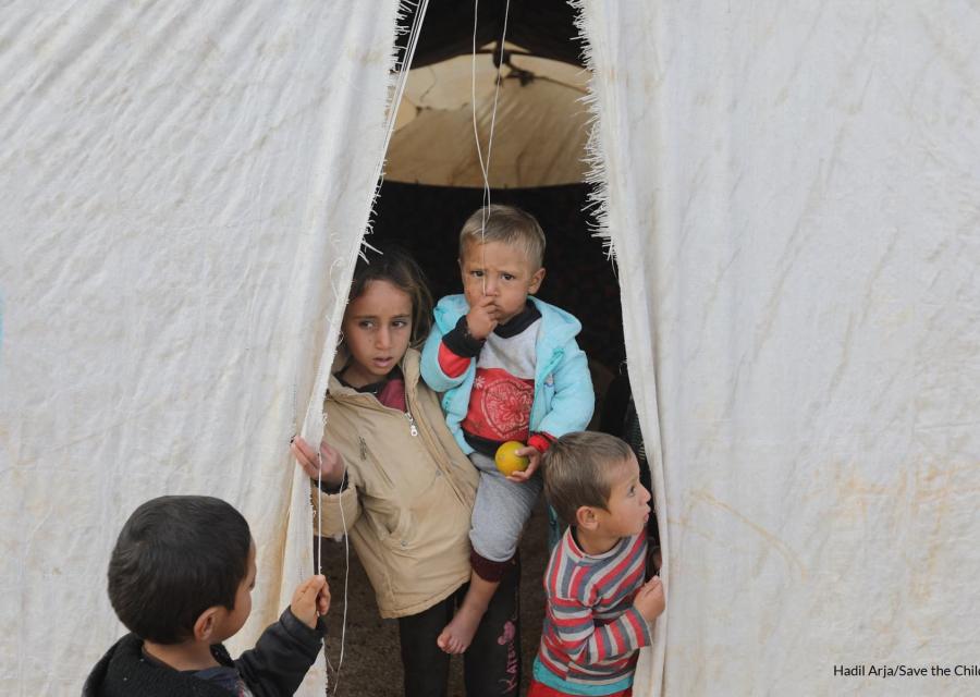 quattro bambini siriani sono sulla soglia di una tenda di un campo rifugiati, una di loro è più grande e tiene in braccio un altro.