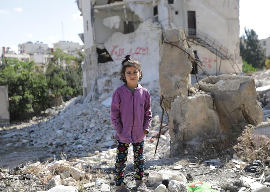 Bambina siriana in piedi tra le macerie. Indossa pantaloni neri e una felpa viola e tiene le mani nelle tasche