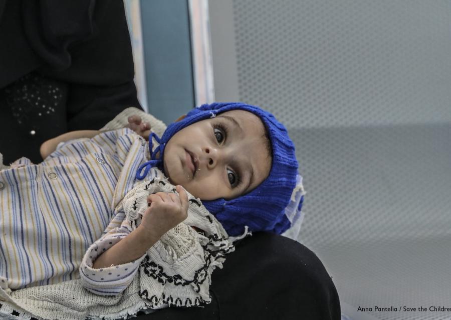 Primo piano di un bambino yemenita di 11 mesi sdraiato sulle gambe della mamma, guarda in alto e indossa un cappello di lana blu e un abitino bianco a righe.