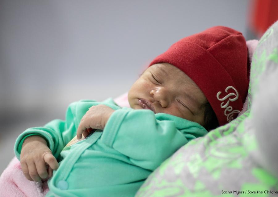 neonato con tutina verde acqua, berrettino rosso, che dorme in braccio alla sua mamma