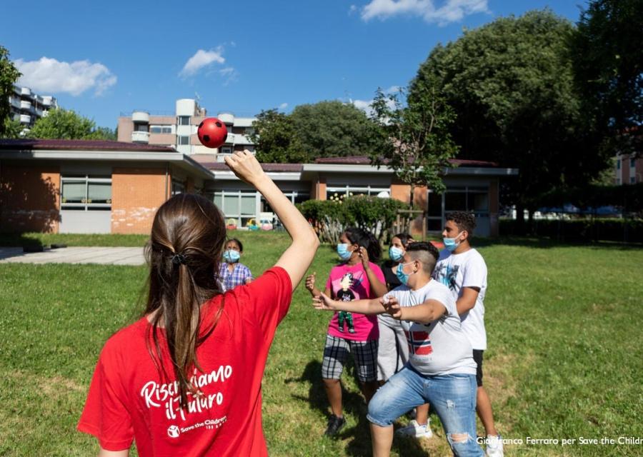 Un operatrice Save the children di spalle gioca a palla con bambini su un prato