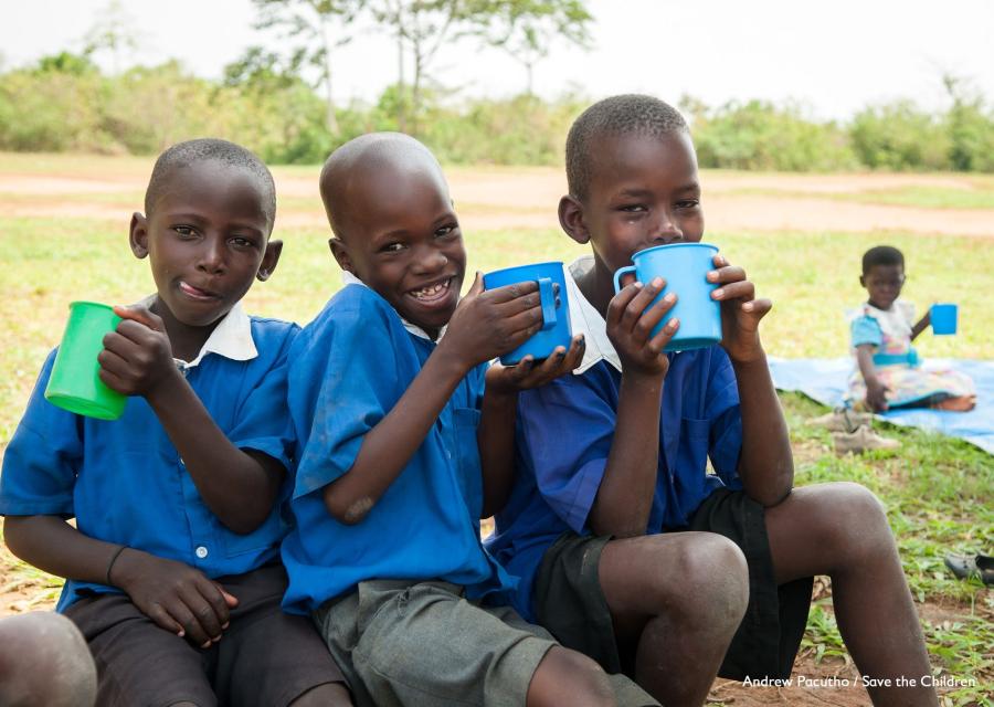 tre bambini ugandesi vestiti di blu, seduti su un ramo bevono acqua da alcune tazze colorate