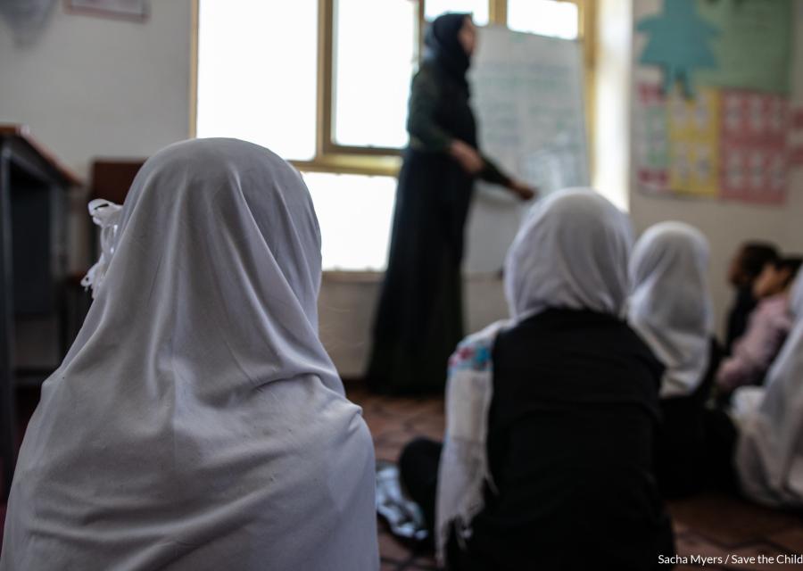 ragazze di spalle, sedute per terra in un aula scolastica, indossano un velo bianco e guardano verso la maestra