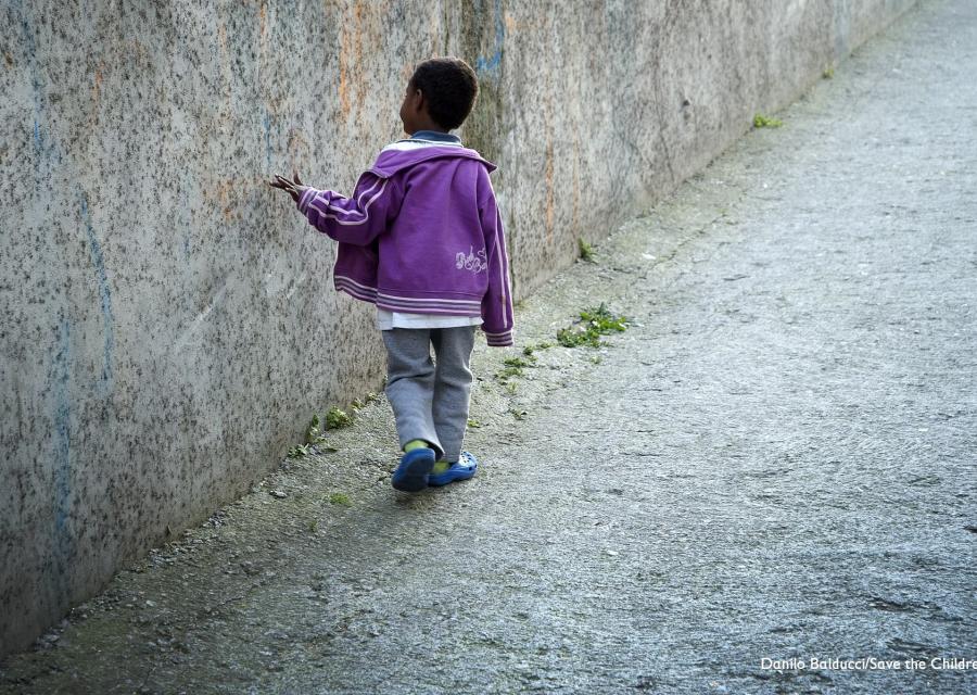 Bambino migrante cammina di spalle toccando muro grigio