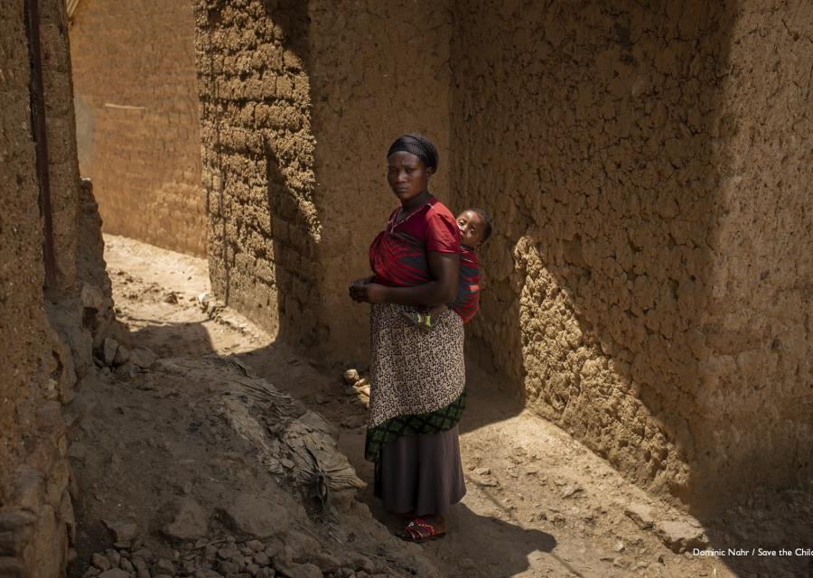 Donna ruandese in piedi tra due mura porta sulle spalle un bambino neonato nella fascia. Lei indossa una maglia rosa e una lunga gonna marrone.