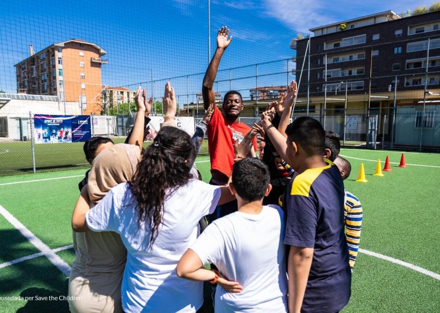 bambini e bambine in campetto da calcio riuniti in cerchio per giocare con operatore Save the Children