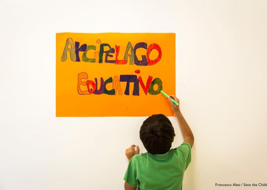 Bambino di spalle con maglietta vede scrive su un cartello appeso al muro le parole arcipelago educativo