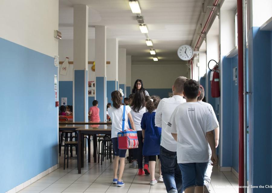 Bambini in fila nel corridoio di una scuola sono inquadrati di spalle mentre camminano verso l insegnate sullo sfondo girata verso di loro