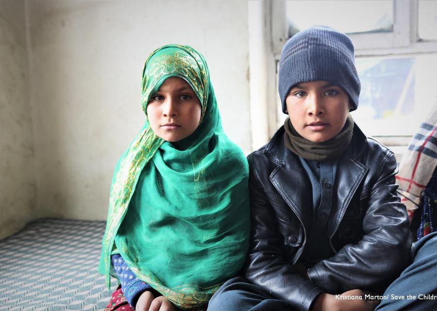 due bambini afghani seduti a terra, un maschio e una femmina. Lei indossa un chador verde e lui un berretto blu e una giacca di pelle nera..