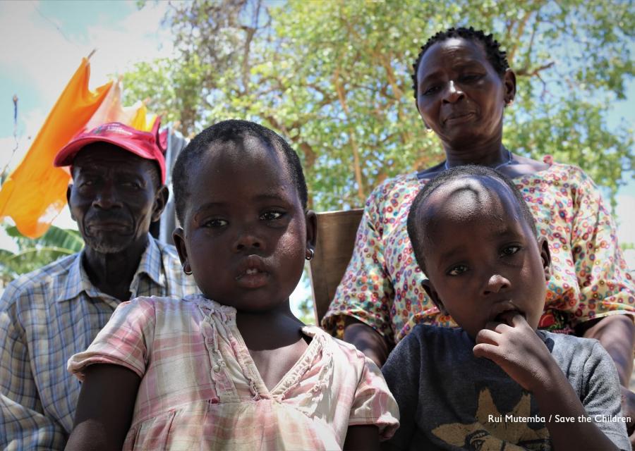 Due bambini del Mozambico in primo piano e dietro di loro un uomo e una donna guardano in camera ripresi leggermente dal basso. Uno dei bambini ha un dito in bocca e una maglietta nera, l altra ha un vestitino bianco