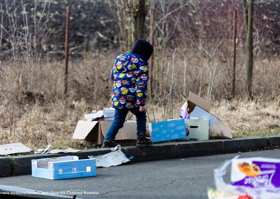 bambino con giacca blu che cammina tra alcune scatole e rifiuti sul ciglio di una strada