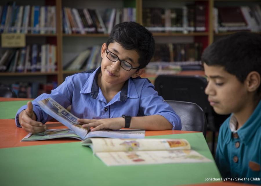 Due bambini seduti al tavolino di una biblioteca leggono i loro libri. Sullo sfondo gli scaffali della biblioteca.