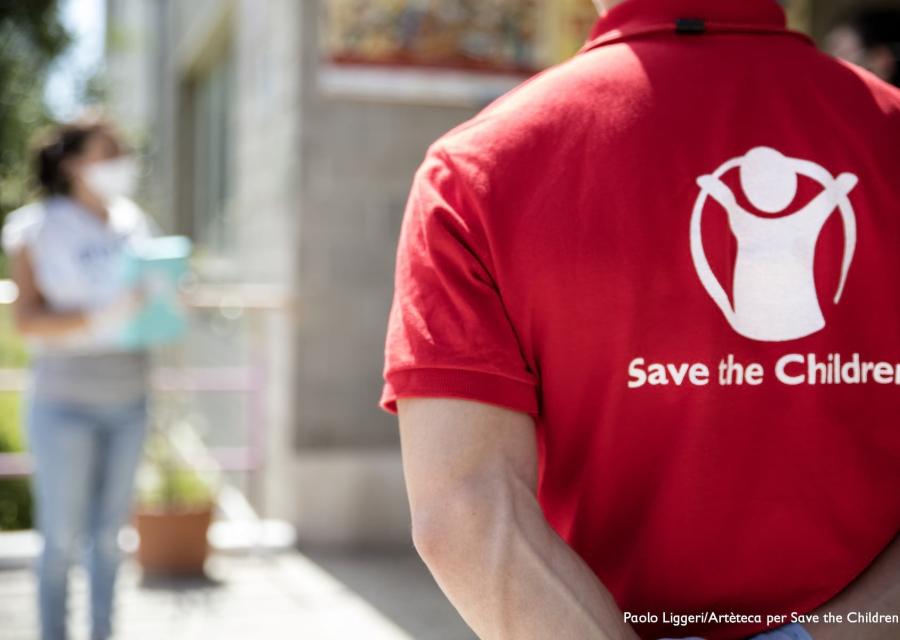 operatore save the children di spalle con maglietta brandizzata in primo piano. Sullo sfondo una figura di donna sfocata
