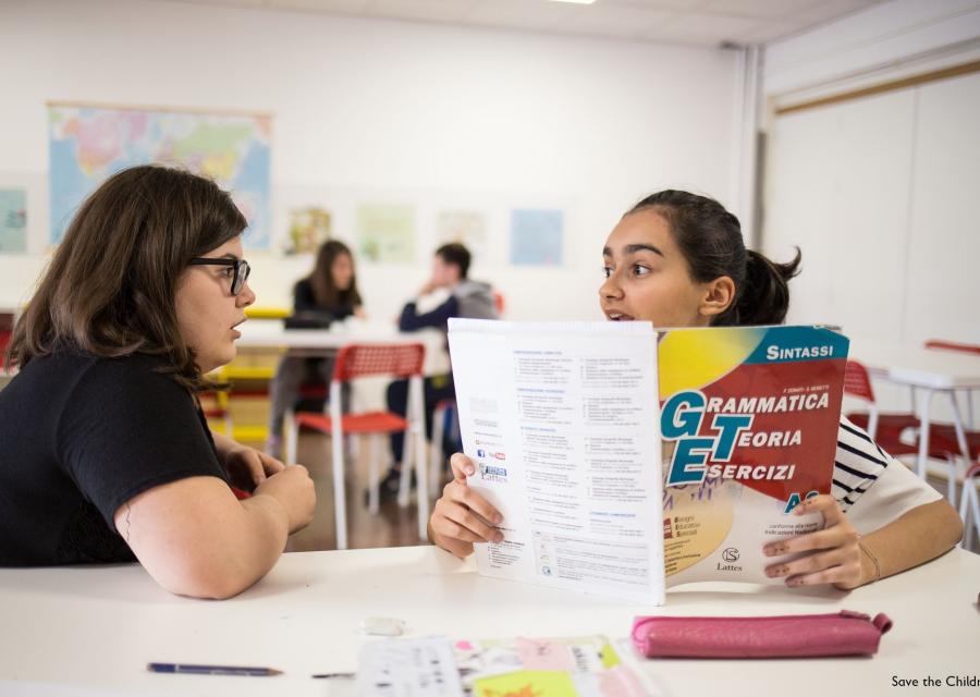 due ragazze sedute al banco di scuola una di loro con un libro di esercizi in mano parlano tra loro