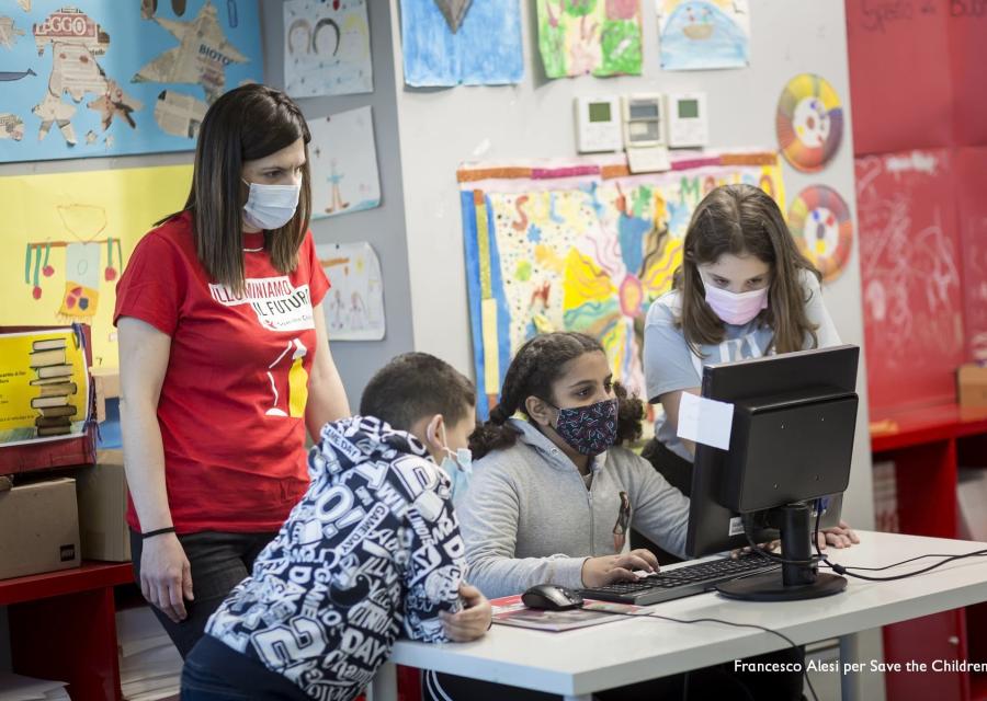 tre bambini e un operatrice save the children lavorano davanti allo schermo di un pc su una scrivania all interno di un Punto Luce