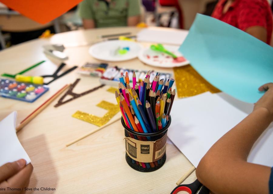 tavolo rotondo con attorno 4 bambini non riconoscibili poiché inquadrati dalle spalle in giù. Sul tavolo materiale da disegno e in primo piano un porta pastelli pieno di pastelli colorati.