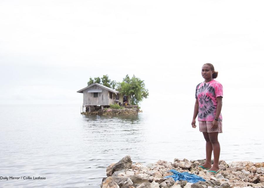 Una ragazza delle Solomon Island è in piedi sulla riva di un lago e la sua casa è un isolotto artificiale alle sue spalle