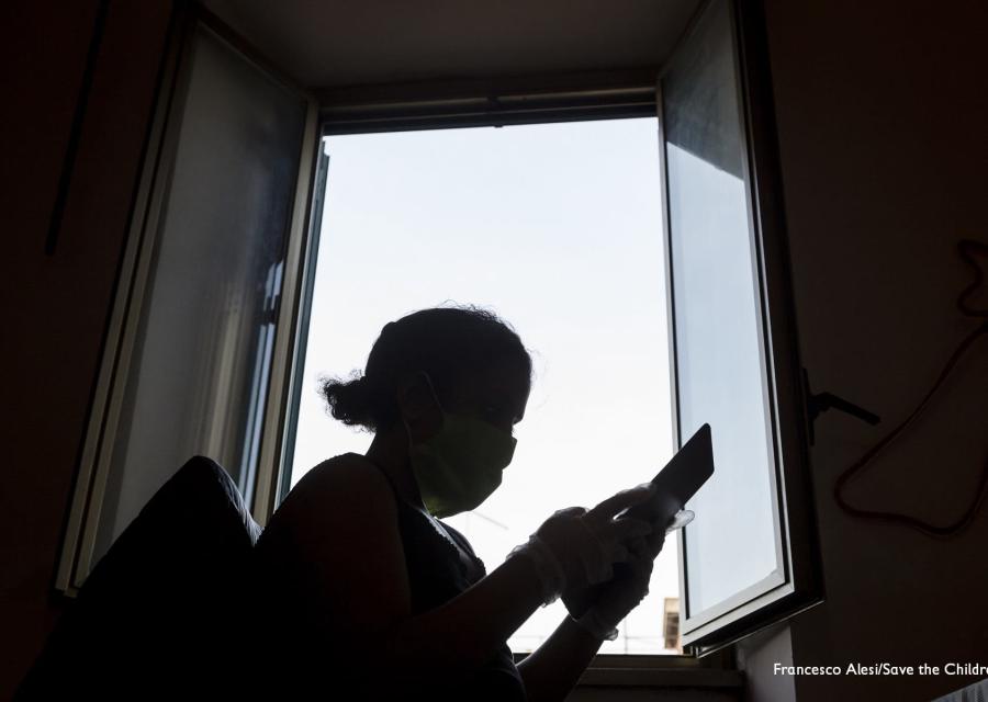 Sagoma nera in controluce di una bambina davanti a una finestra che tiene in mano un tablet e ne guarda lo schermo