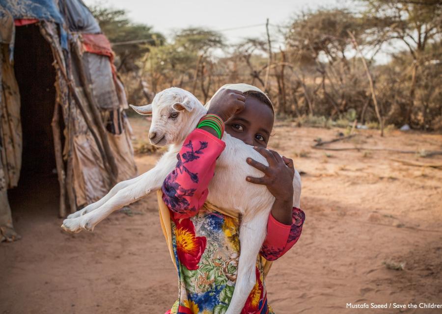 bimba somala con belo bianco e vestito lungo colorato con in braccio una capretta bianca fuori dalla sua abitazione in una zona semi desertica 