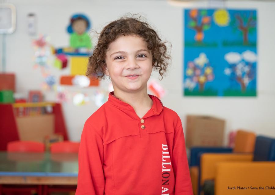 bambina sorridente con felpa rossa guarda in camera. Si trova in una scuola per l'infanzia.