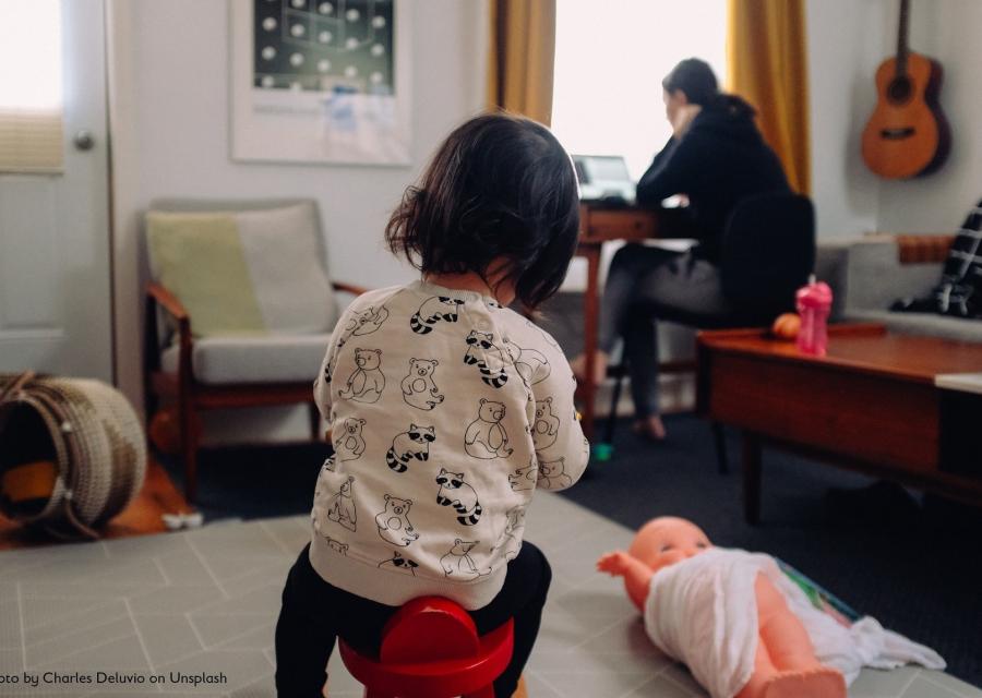 bimba in primo piano di spalle che gioca in casa e sullo sfondo la mamma che lavora al pc