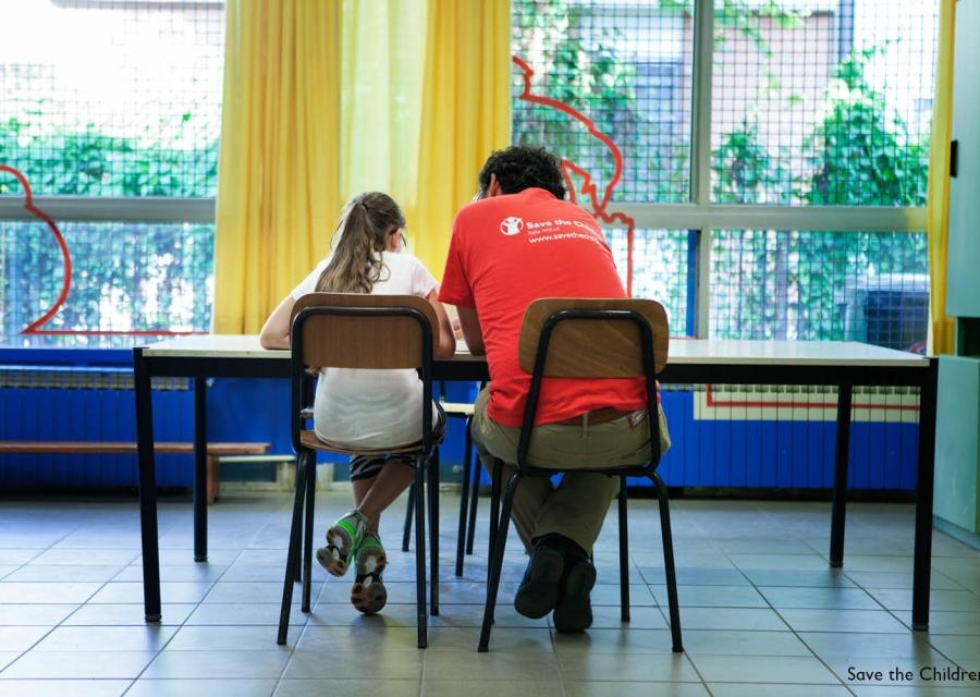 operatore save the children di spalle seduto a un tavolo con affianco una bambina seduta
