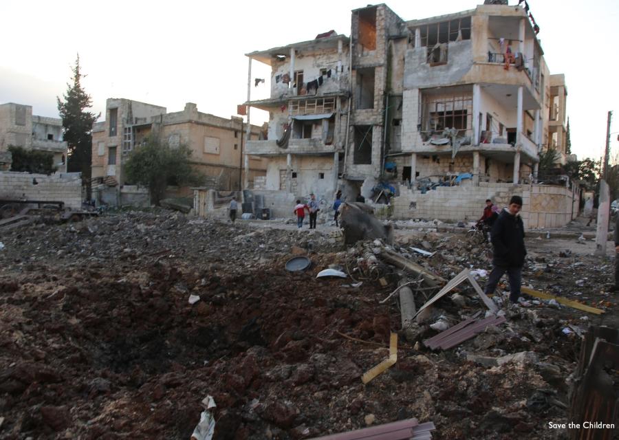Siria, macerie di palazzi e in sfondo un palazzo semi distrutto dai bombardamenti. Tra le macerie camminano alcuni adulti e bambini.