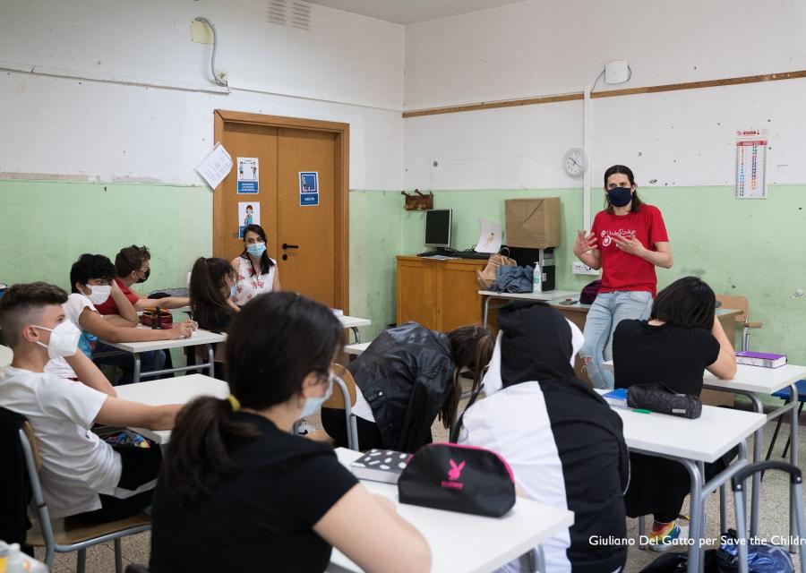 classe di studenti ripresi da dietra seduti ai banchi, in fondo un insegnante in piedi appoggiato alla cattedra spiega