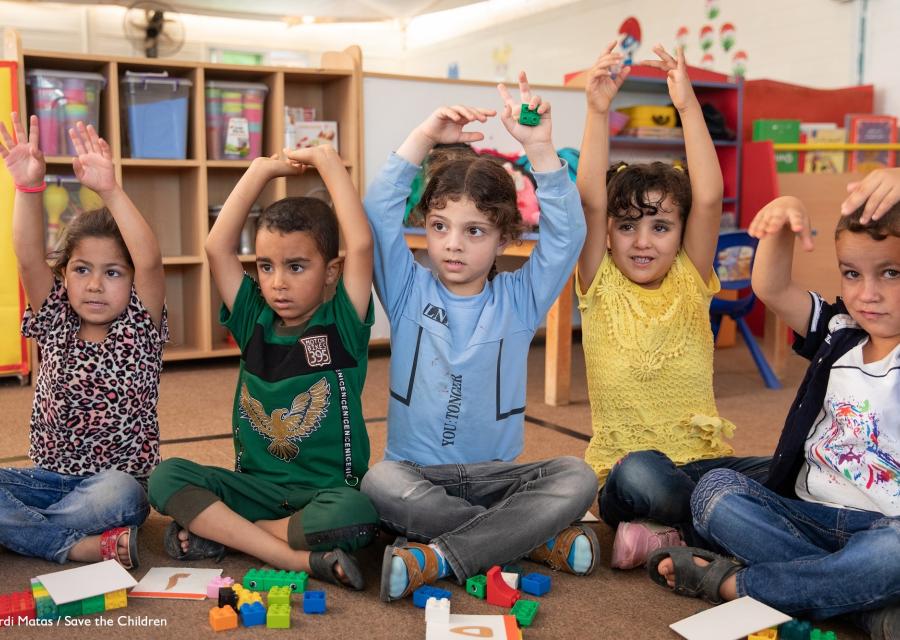 5 bambini seduti in cerchio con le mani alzate durante una lezione a scuola