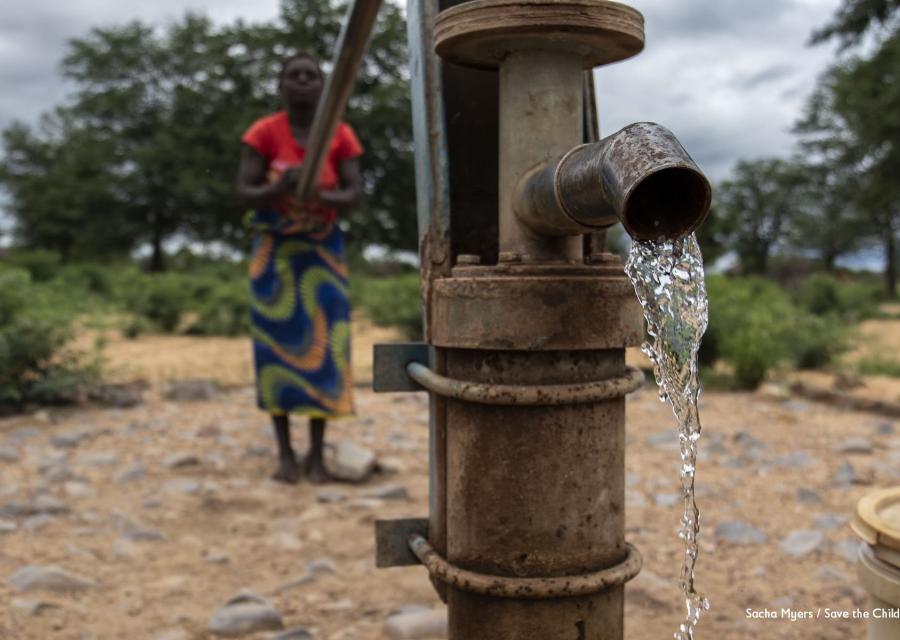 In primo piano una fontana a pompa da cui sgorga acqua, dietro sfocata si vede una donna che aziona la pompa per far uscire l acqua