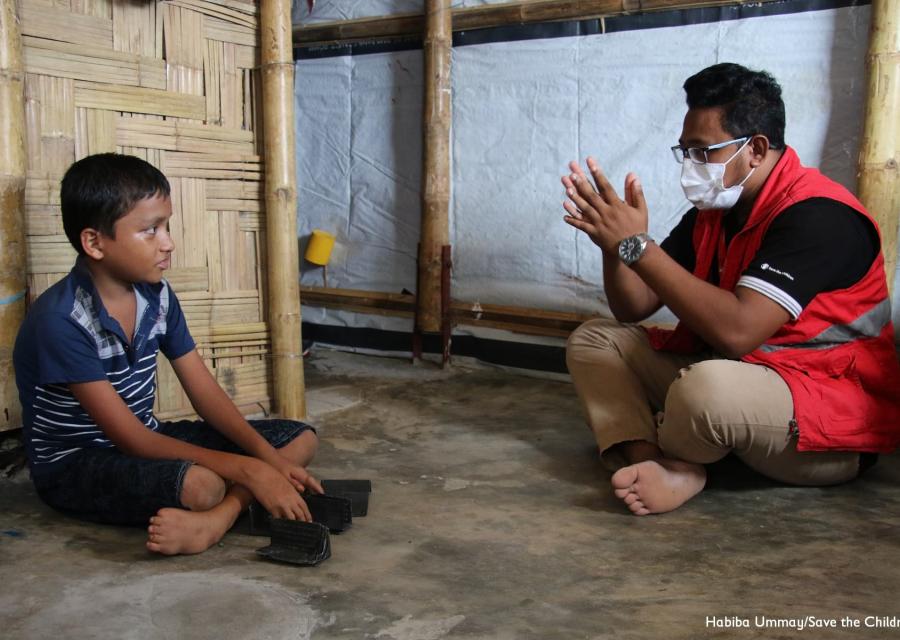 Un bambino rohingya è seduto a terra a gambe incrociate  e davanti a lui un operatore di save the children gesticola con le mani.