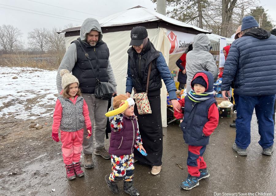 Una famiglia con due genitori e tre bambini si tengono per mano in strada. Hanno vestiti invernali molto pesanti e si vedono fiocchi di neve che cadono dal cielo. Anche dietro di loro il prato è ricoperto di neve.