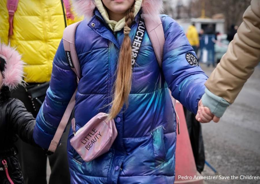 una bambina al centro della scena viene ripresa senza la testa mentre cammina tenendo la mano a due persone che le sono a fianco. Indossa un giubbotto blu e una borsetta rosa a tracolla.