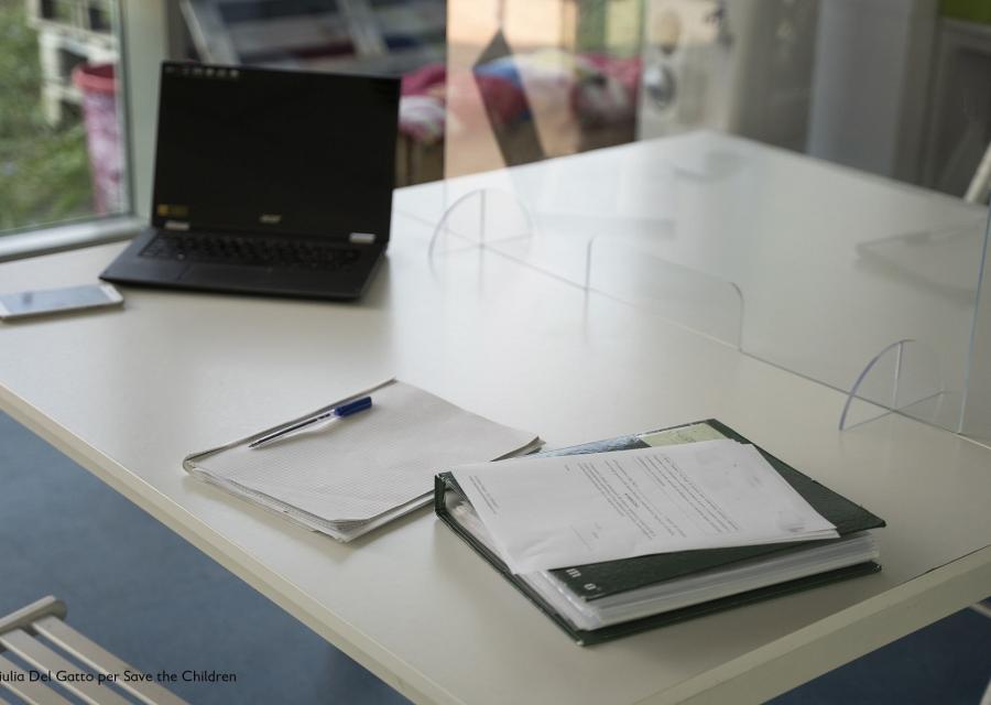 tavolino bianco con divisorio in plexiglass con sopra schedario, penna, un lap top e alcuni fogli