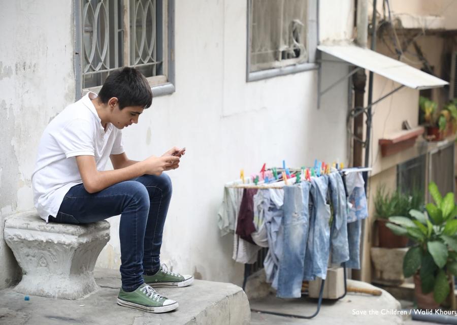 Un ragazzo adolescente  seduto su un sedile di marmo è piegato a guardare il suo cellulare. Indossa un paio di jeans, scarpe verdi e una maglietta bianca. Accanto a lui uno stendino con i panni stesi.