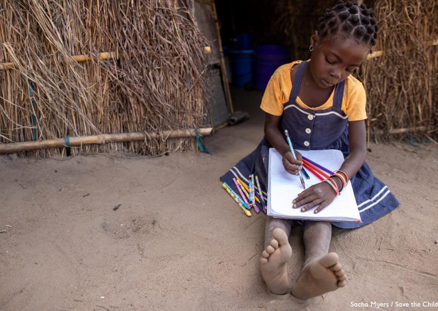 una bambina africana con maglietta gialla e vestito jeans è seduta a terra accanto alla sua capanna mentre è intenta a scrivere qualcosa sopra un quaderno