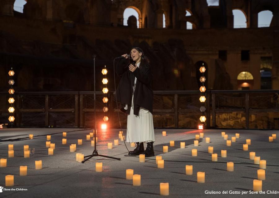 La cantautrice Elisa canta in piedi con microfono all interno del Colosseo circondata da candele accese sul palco. Elisa indossa un vestito lungo bianco e una giacca lunga nera