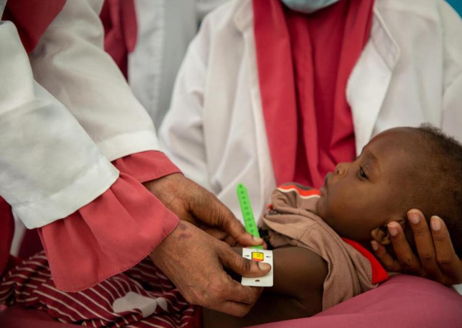 bambino sorretto da due operatrici mentre una gli misura il livello di malnutrizione con un braccialetto muac