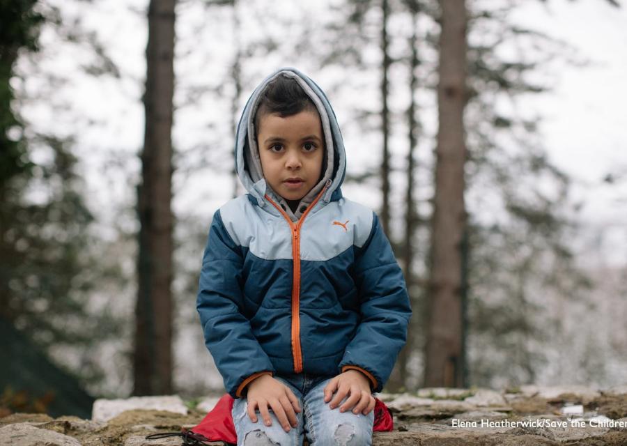Bambino seduto su un muretto e sguardo in camera con sfondo bosco innevato