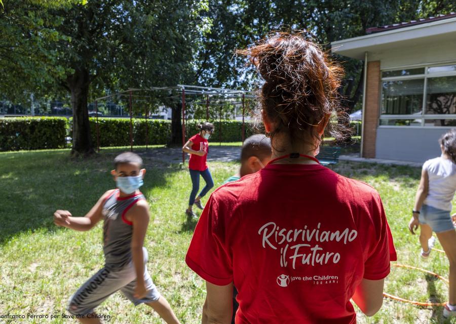 ragazza di spalle con maglietta rossa insieme a dei bambini che giocano nel prato con indosso la mascherina