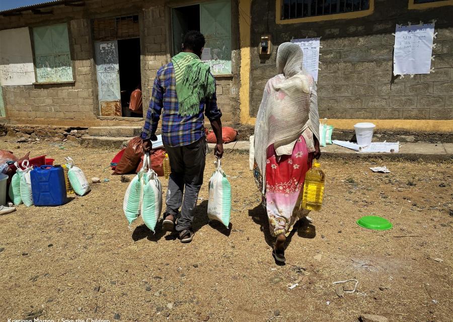 due persone di spalle che camminano verso una casa su un terreno sterrato portando con sè acqua e cibo